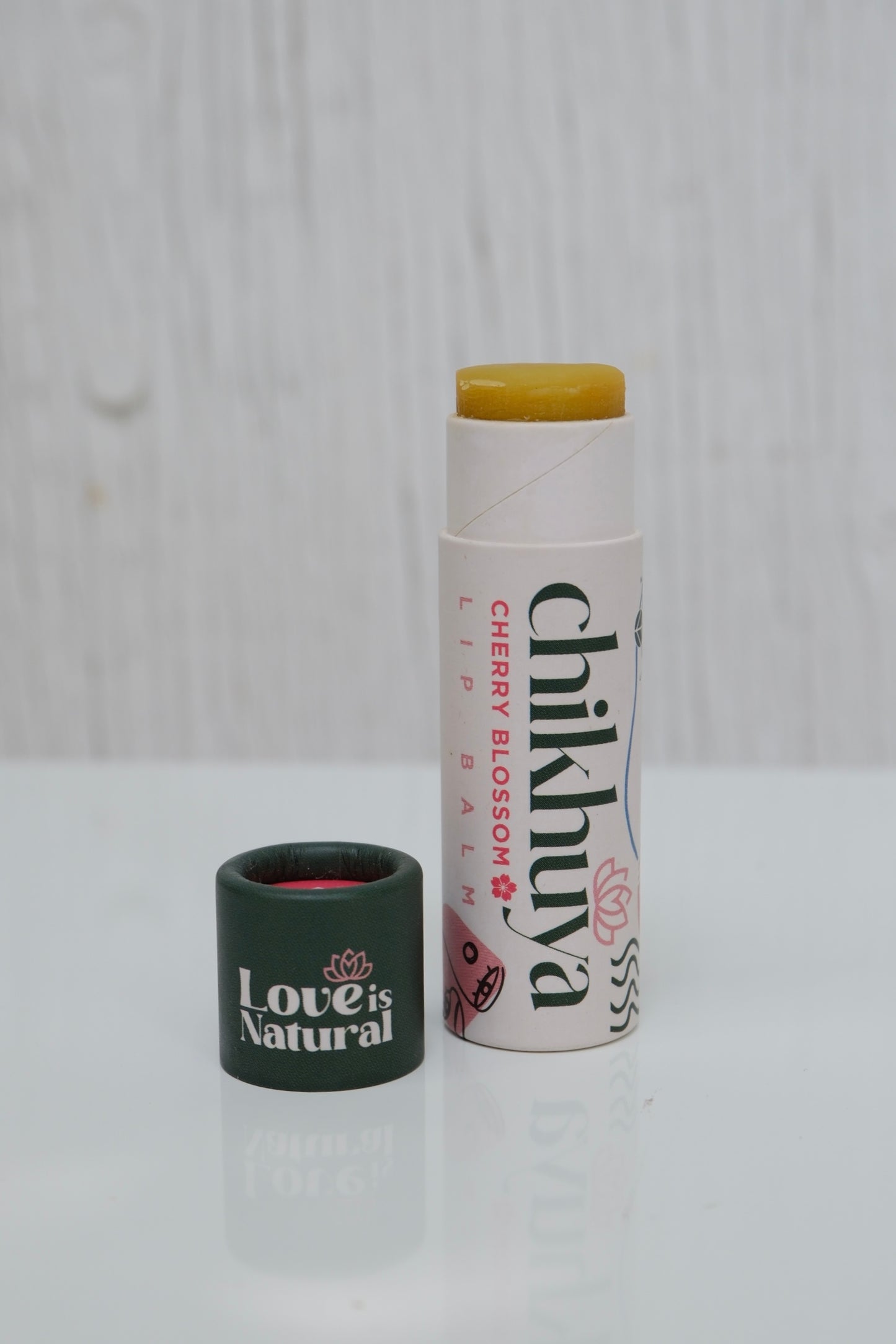 Branded lip balm in a white stick and green cap of the balm next to it with 'Love is Natural' text. Product is on a light gray background.