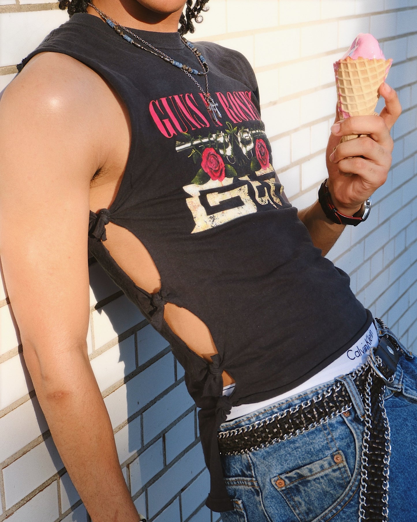 Man wearing a black tank top with 'Guns N' Roses' graphic design and ties on the side, holding an ice cream cone against a white brick wall