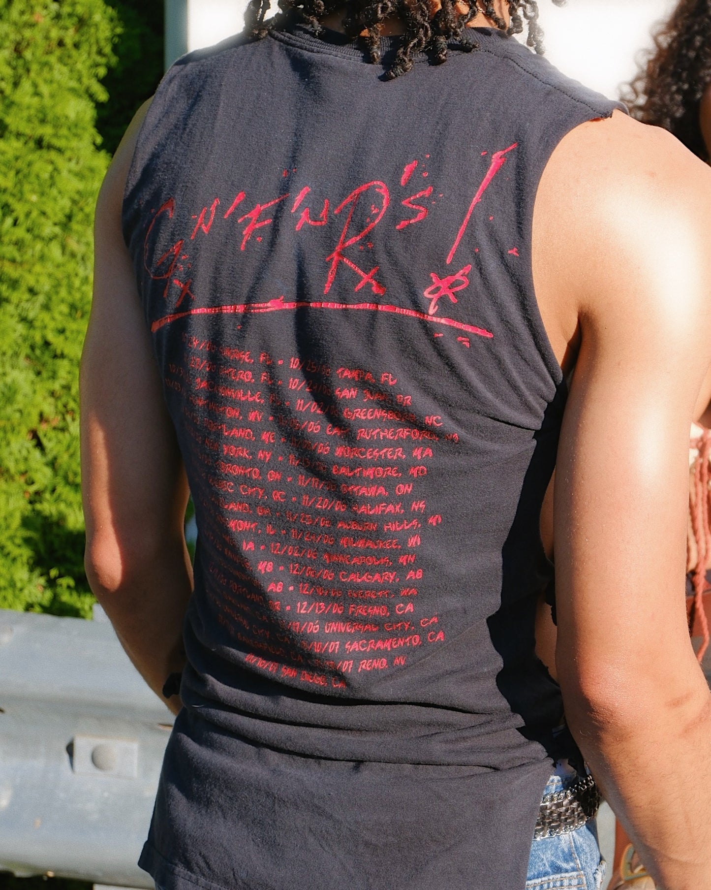 Man is standing outside wearing a 'Guns N' Roses' tank top with tour dates listed on the back in red text