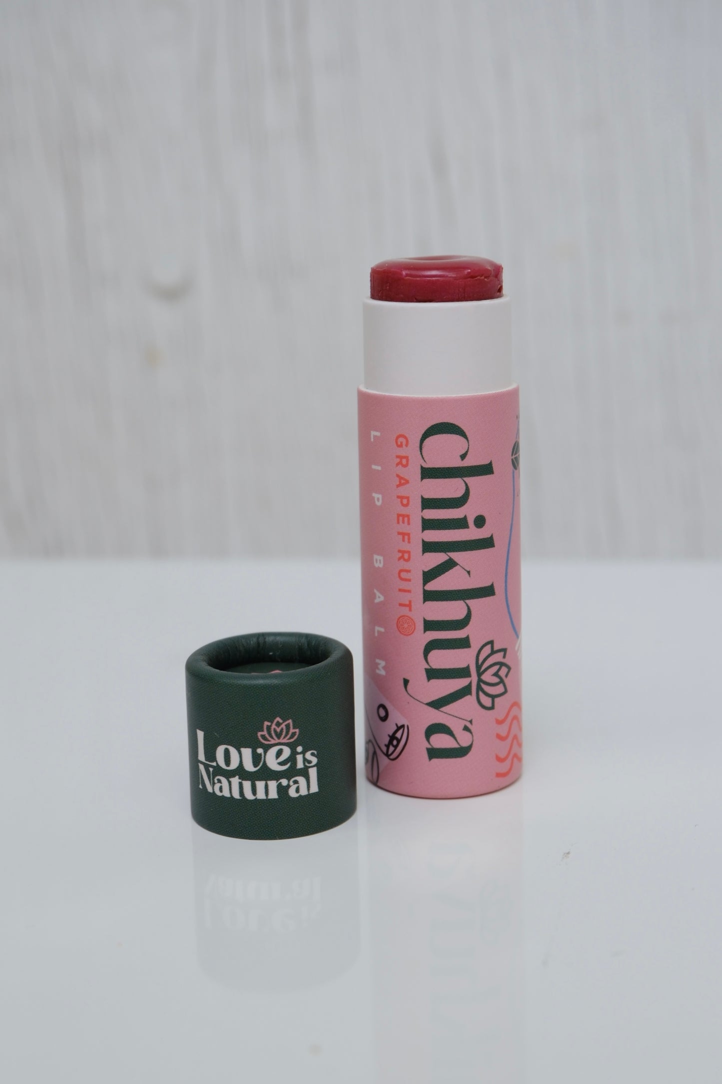 Branded lip balm in pink stick and green cap of the balm next to it with 'Love is Natural' text. Product is on a light gray background.