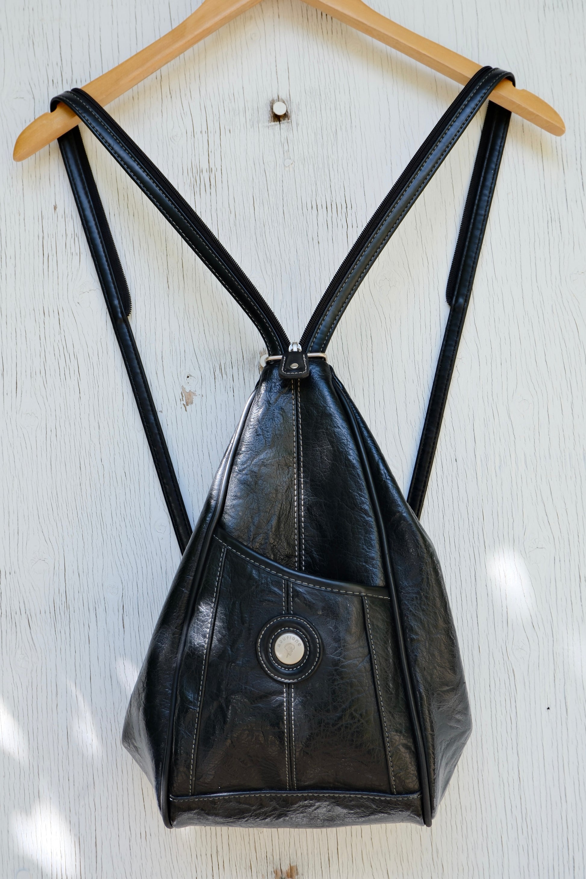 Black faux leather bag, with strap unzipped as a backpack, on a hanger against a light wooden background
