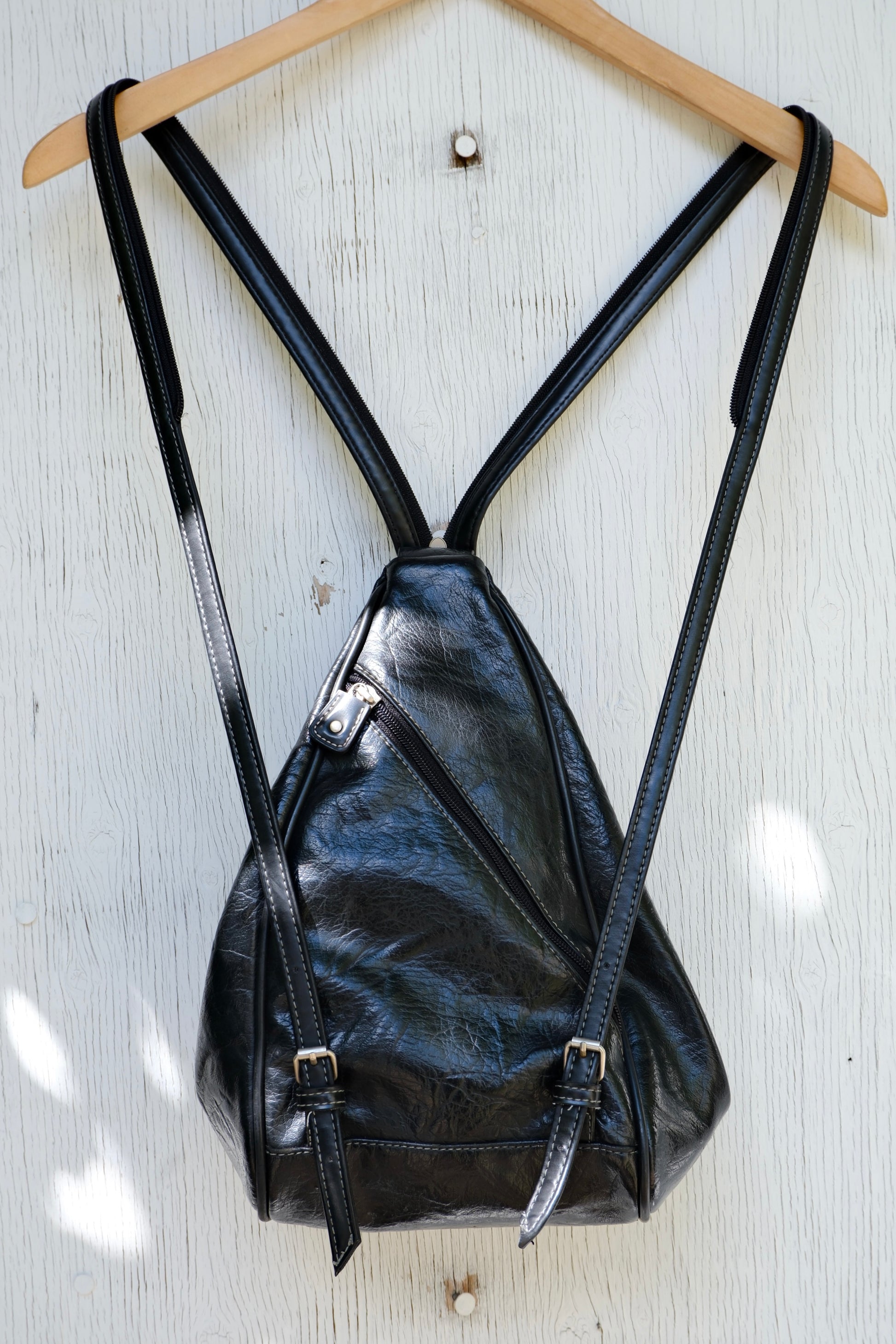 Back of the black faux leather bag, with strap unzipped as a backpack, on a hanger against a light wooden background